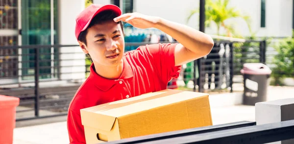 Happy delivery man in red uniform holding cardboard box standing near house of customer.courier service concept