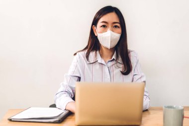 Koronavirüs için karantinada bilgisayar kullanan Asyalı genç iş kadını ofis masasında otururken koruyucu maske takıyor.