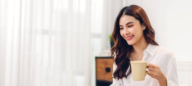 Gülümseyen mutlu, neşeli, güzel Asyalı bir kadının portresi rahatlıyor, içiyor ve bir fincan sıcak kahve ya da çay seyrediyor.