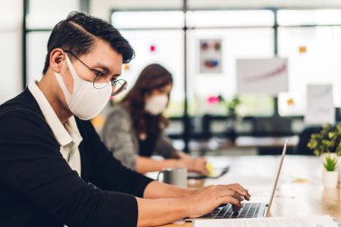 Koronavirüs için karantinada bilgisayar kullanan Asyalı iş adamı ve kadın ofis masasında otururken koruyucu maske takıyor ve bilgisayar kullanıyor.