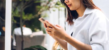 Gülümseyen mutlu Asyalı kadın portresi dijital teknoloji akıllı telefon kullanarak rahatlıyor. Genç Asyalı kız ekranda yazı yazma ve kafede oyun oynama veya sosyal medya oyunlarına bakıyor.