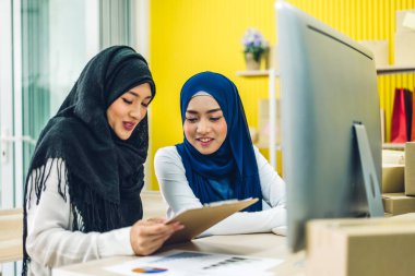 Müslüman, iki Asyalı serbest çalışan kadın internetten alışveriş yapıyor ve karton kutuları evde, iş yeri online nakliye ve teslimat konseptiyle kontrol listesi düzenliyor.