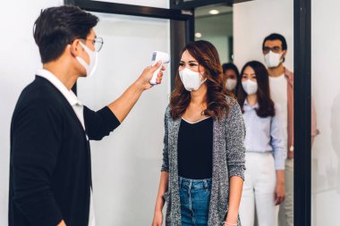 Bir grup profesyonel Asya işi ve ofiste koruyucu maske takarak koronavirüs için karantinada çalışmadan önce vücut ısısını ölçmek için kızılötesi termometre kullanılıyor.