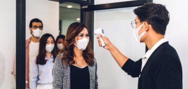 Bir grup profesyonel Asya işi ve ofiste koruyucu maske takarak koronavirüs için karantinada çalışmadan önce vücut ısısını ölçmek için kızılötesi termometre kullanılıyor.