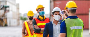 Koronavirüs için karantinada çalışırken koruyucu maske takıp vücut ısısını ölçmek için kızılötesi termometre kullanan kasklı grup şefi.