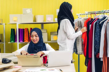 Müslüman, iki Asyalı serbest çalışan kadın internetten alışveriş yapıyor ve karton kutuları evde, iş yeri online nakliye ve teslimat konseptiyle kontrol listesi düzenliyor.