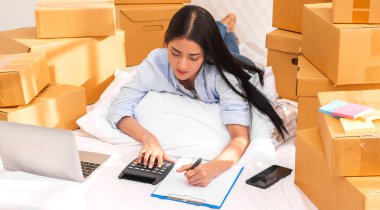 Genç, gülümseyen güzellik sahibi Asyalı kadın serbest çalışan bir iş adamı dizüstü bilgisayarla alışveriş yapıyor evde yatağında paket kutusuyla çalışıyor - KOBİ işi online ve teslimat konsepti