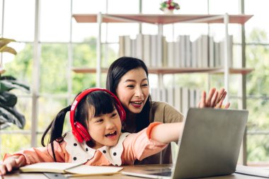 Anne ve Asyalı küçük kız öğreniyor ve dizüstü bilgisayara bakıyor çevrimiçi eğitim e-öğrenim sistemiyle ödev yapıyor. Evde öğretmenle çocuk video konferansı.