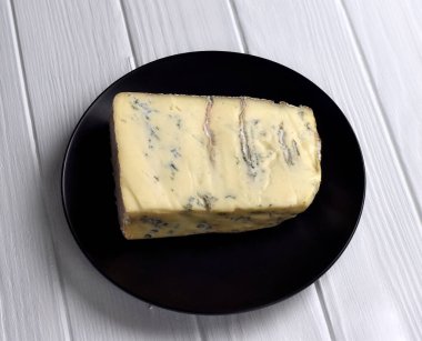Bir dilim mavi damarlı Gorgonzola peyniri beyaz ahşap masada siyah bir tabakta..