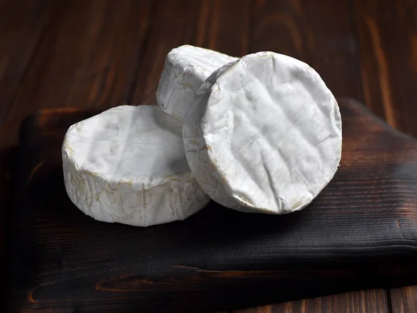 Three wheels of Camembert soft cheese with white mold on a dark wooden cutting board.