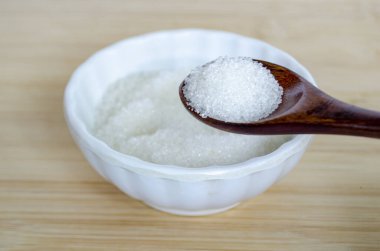 Beyaz kâsenin üzerine tahta kaşıkla şeker tanelerinin yakın ayrıntıları glikoz kontrolünü ve önleyici sağlık stratejisini yansıtıyor.