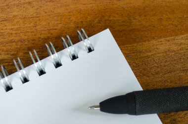 Tahta bir masanın üzerinde boş bir spiral defter, alt tarafında siyah bir kalem, metin ya da fikir için özelleştirilebilir bir not defteri var.