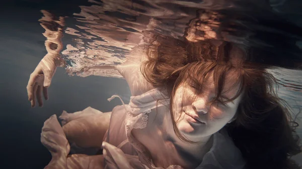 Girl in a dress underwater as if in zero gravity, light blue background and beautiful reflection on the water surface