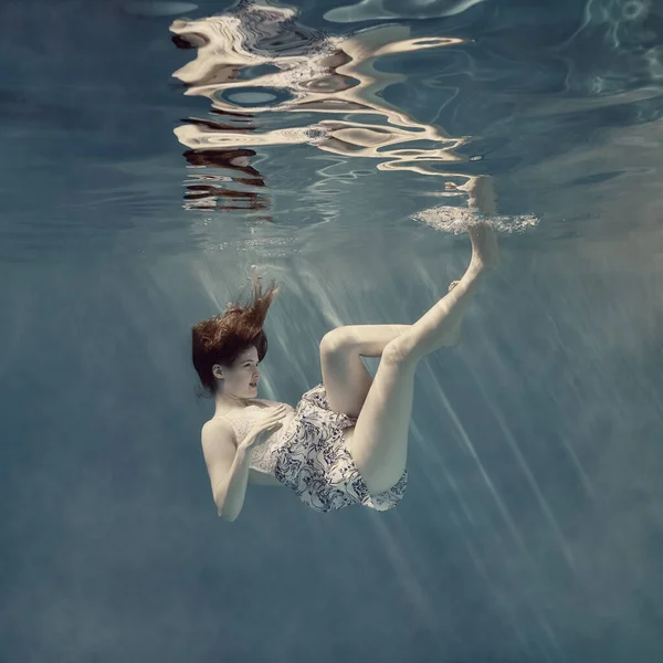 Girl in a dress underwater as if in zero gravity, light blue background and beautiful reflection on the water surface