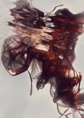 Kurdeleli ve havadar kumaşlı kırmızı elbiseli bir kadın, sanki sıfır yerçekimindeymiş gibi arka planda su altında poz veriyor..