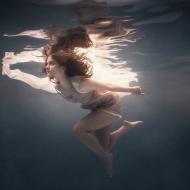 Uzun siyah saçlı bir kız, karanlık bir arka planda suyun altında yüzer, sanki ağırlıksız yüzer gibi.