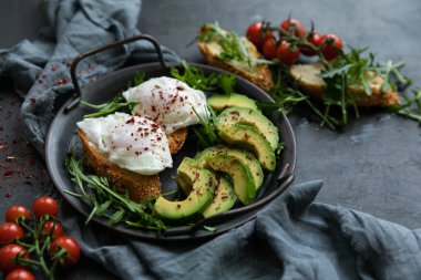 Bir dilim kızarmış ekmek, avokado, roka, vişneli domates ve baharatla nefis ve lezzetli bir kahvaltı. Dökme demir bir tavada servis edilmiş, siyah arka planda tekstil ile servis edilmiş. .