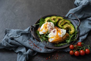  Bir dilim kızarmış ekmek, avokado, roka, vişneli domates ve baharatla nefis ve lezzetli bir kahvaltı. Dökme demir bir tavada servis edilmiş, siyah arka planda tekstil ile servis edilmiş. .