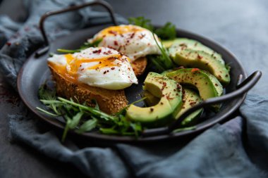 Bir dilim kızarmış ekmek, avokado, roka, vişneli domates ve baharatla nefis ve lezzetli bir kahvaltı. Dökme demir bir tavada servis edilmiş, siyah arka planda tekstil ile servis edilmiş. .