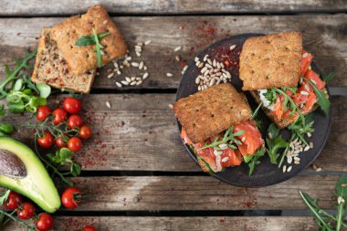 Öğle yemeği için lezzetli bir aperatif. Hafif tuzlu somonlu lezzetli bir sandviç. Tüylü bir çörek, arkaplanda roka, ayçekirdeği, vişneli domates ve avokado..