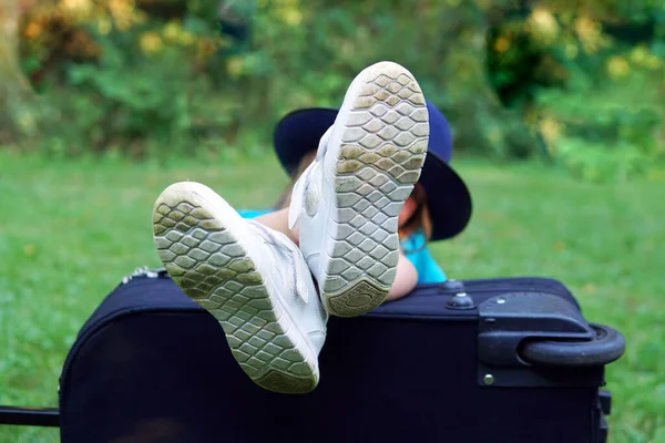 sneakers feet rest a girl in a hat with a suitcase is sitting on the grass going on a trip