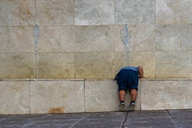 Mavi şortlu, sırtı tanınmayan bir çocuk çömelmiş mermer bir korkulukta oynuyor.