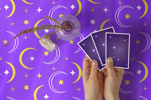 Mor yıldızlar ve hilal desenli masa örtüsü üzerinde üç tarot ya da kahin kartı tutan altın ojeli genç bir kadının elleri.