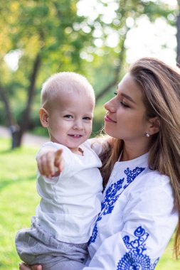 Anne, güneşli bir günde beyaz elbiseli oğluyla parkta el ele tutuşup oynaşıyor ve sarılıyor.