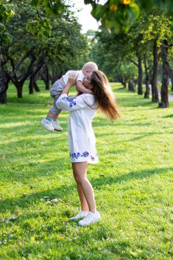 Anne, güneşli bir günde beyaz elbiseli oğluyla parkta el ele tutuşup oynaşıyor ve sarılıyor.