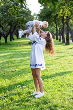 Anne, güneşli bir günde beyaz elbiseli oğluyla parkta el ele tutuşup oynaşıyor ve sarılıyor.