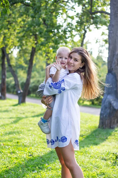 Anne, güneşli bir günde beyaz elbiseli oğluyla parkta el ele tutuşup oynaşıyor ve sarılıyor.