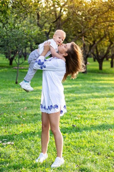 Anne, güneşli bir günde beyaz elbiseli oğluyla parkta el ele tutuşup oynaşıyor ve sarılıyor.