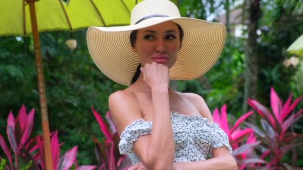 Beautiful girl in a hat looking at the camera, tropical flowers in background — Stock Video