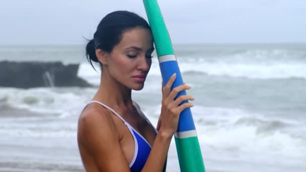 Chica está de pie con una tabla de surf contra el fondo de grandes olas del océano — Vídeos de Stock