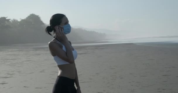 Maskeli güzel kız yazın kumlu sahilde uzanıyor ve müzik dinliyor. — Stok video