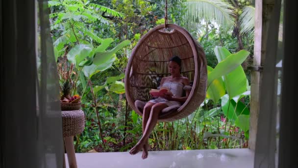 A girl sits in a hanging hammock, with a book ,Bali — Stock Video