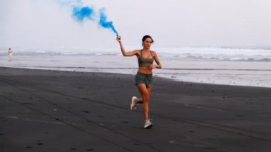 Siyah kumsalda koşan spor kızı. Mavi renkli dumanla, Maraton.