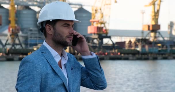 Um homem em um capacete protetor no porto de carga do mar fala no telefone, EUA — Vídeo de Stock