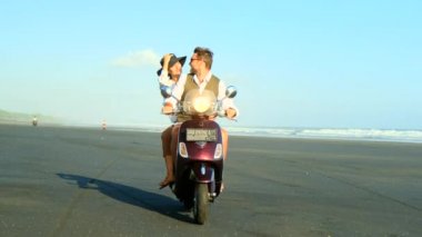 Güzel bir çift okyanus kıyısında koşar, geriye dönük scooter kumda durur.