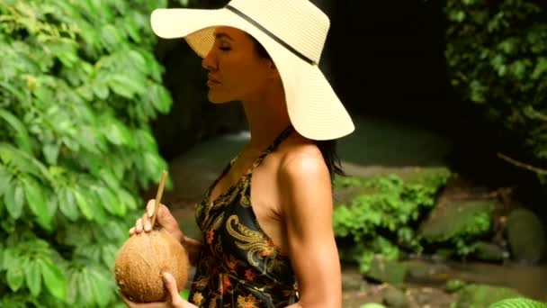 Chica de verano en traje de baño con bebida de coco en la naturaleza tropical cerca de watefall — Vídeo de stock