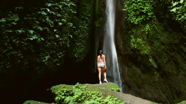 Sexig kvinna står på sten på det vackra vattenfallet i djungeln bredvid klippan — Stockvideo