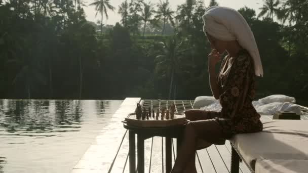 Menina sexy senta-se em um roupão de banho perto da piscina em uma floresta tropical e joga xadrez — Vídeo de Stock