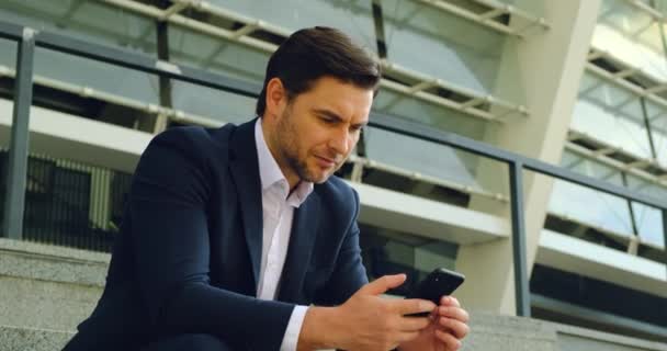 Jóképű üzletember kezében mobiltelefon ül a lépcsőn, közel az irodához — Stock videók