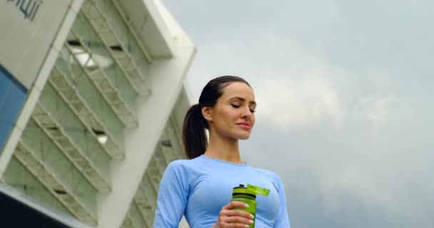 Спортсменка в модной одежде пьет воду после тренировки. Жажда, обезвоживание — стоковое видео