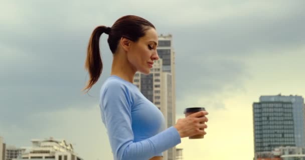 Счастливая спортивная женщина расслабляется после долгой поездки, пьет кофе возле стадиона — стоковое видео