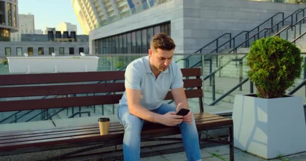 Joven hombre de negocios barbudo sentado en el banco en el parque, mensajes de texto en el teléfono celular — Vídeo de stock