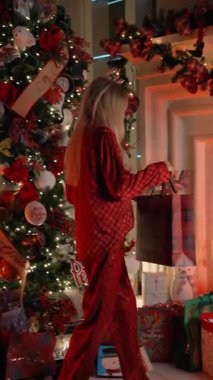 Bayramlık pijamalı bir kadın Noel ağacının altına şöminenin yanına hediye koyar. Dişi, sıcak atmosferi, dekorasyonu ve parlayan ışıklarıyla bayram havası yaratır. Sahne neşeyi, geleneği ve aileyi simgeler.