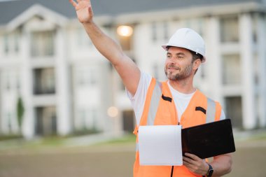 Güvenlik kasklı inşaat yöneticisi parmak kaldırıyor, klasörü ve evrakları açık tutuyor, proje detaylarını gözden geçiriyor. Erkek mühendis yerleşim yerinde. Odaklanmış yönetim atmosferi, modern konut