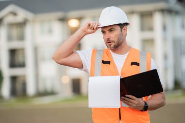 Güvenlik kasklı inşaat yöneticisi parmak kaldırıyor, klasörü ve evrakları açık tutuyor, proje detaylarını gözden geçiriyor. Erkek mühendis yerleşim yerinde. Odaklanmış yönetim atmosferi, modern konut