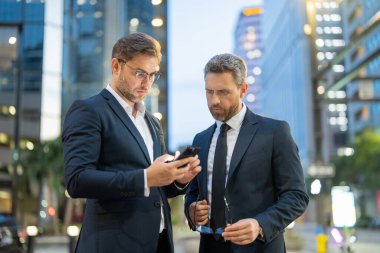 Girişimciler modern bir şehirde dururken akıllı telefondaki bilgileri analiz ederler. Resmi takım elbiseli iki erkek işadamı odaklanma ve konsantrasyonla stratejiyi tartışıyor. Sahne hırs ve takım çalışmasını gösteriyor